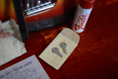 Two keys on a packet with a can of Fender polish and a polishing cloth on a red surface.