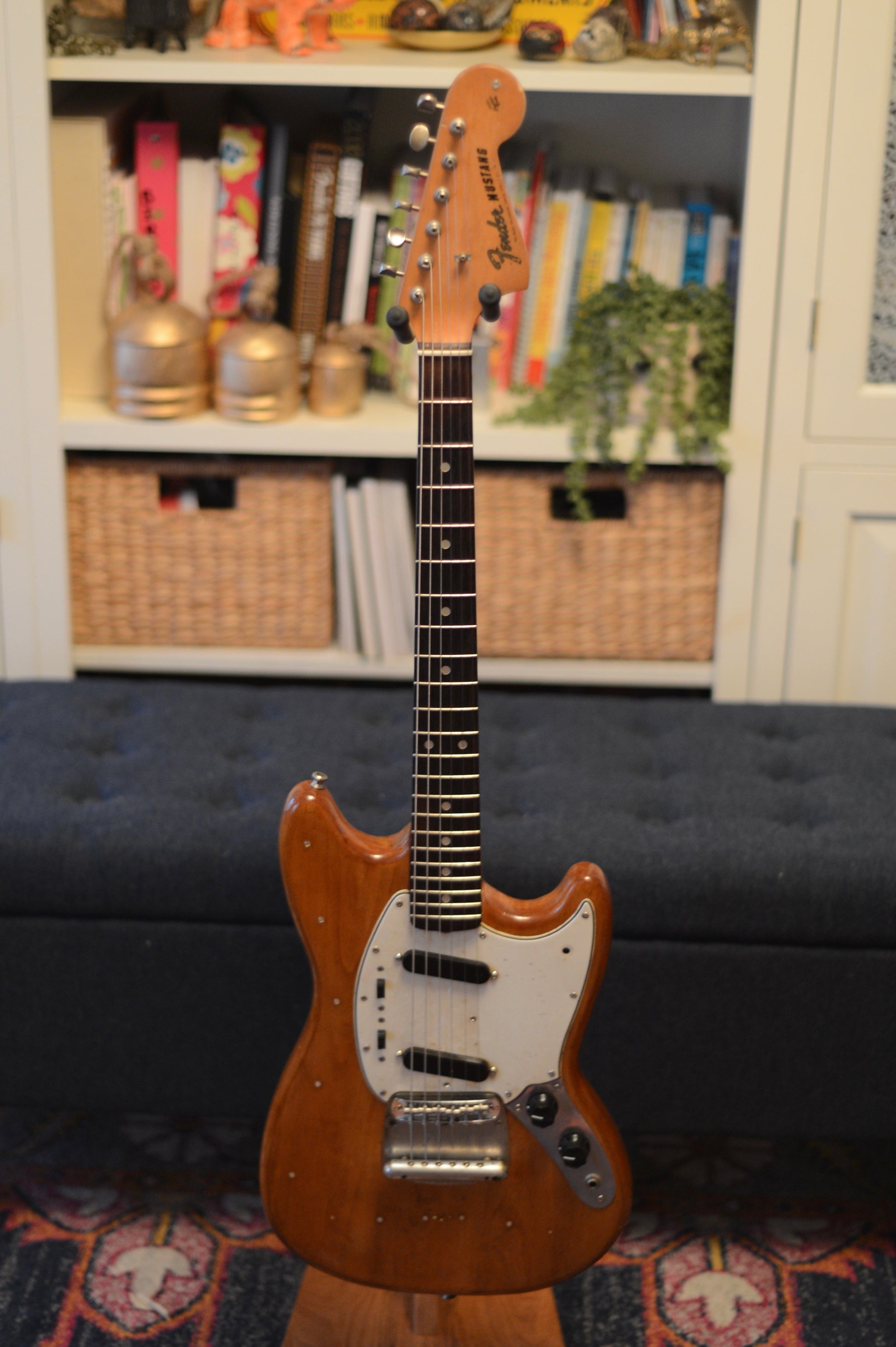 1965 Fender Mustang with a natural finish on a stand in a room with shelves and a couch.
