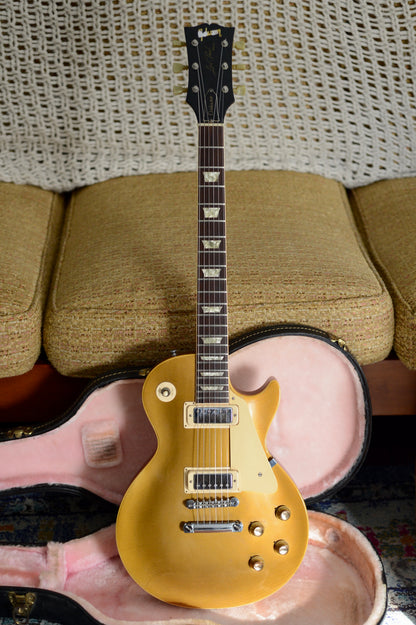 Gold electric Les Paul Deluxe guitar on a pink cushion with a textured wall background